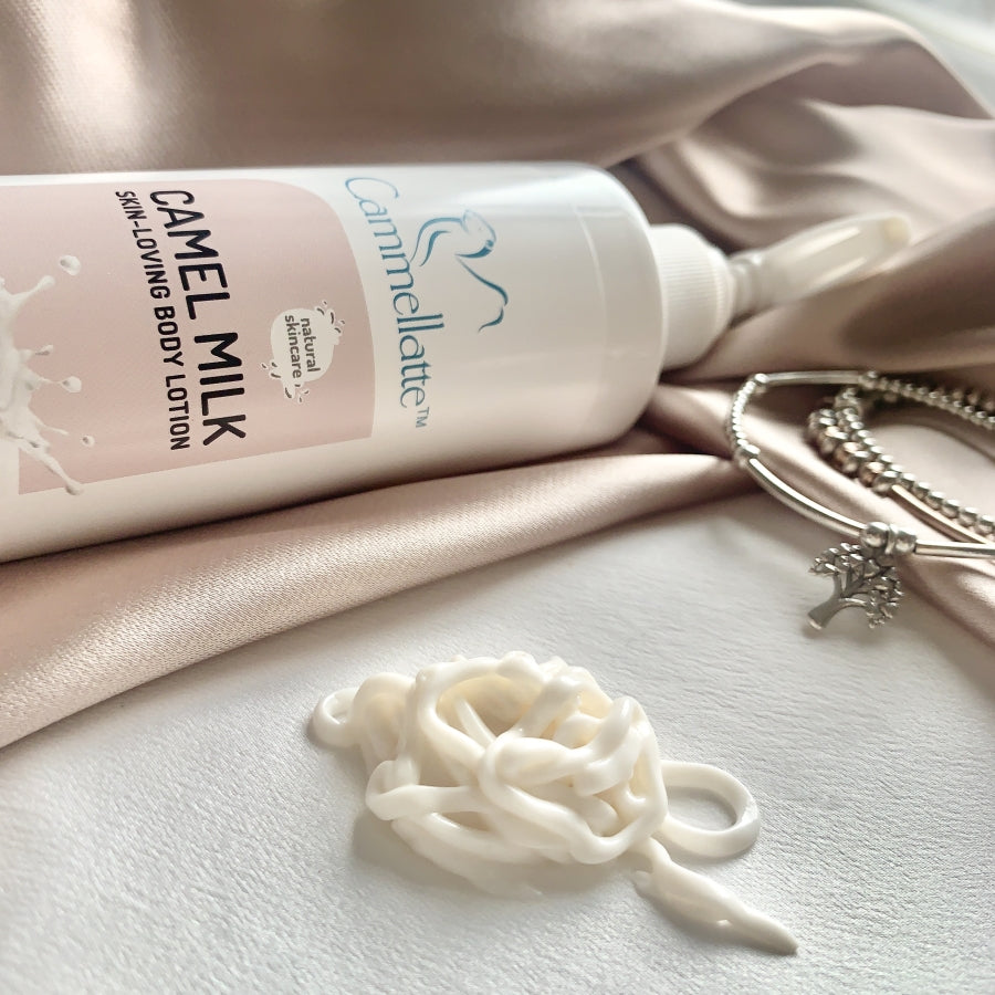 A bottle of Cammellatte's Camel Milk Body Lotion sits elegantly on a silky fabric, with a swirl of the soothing cream beside it. Nearby, silver bracelets glisten next to a small Tree charm.