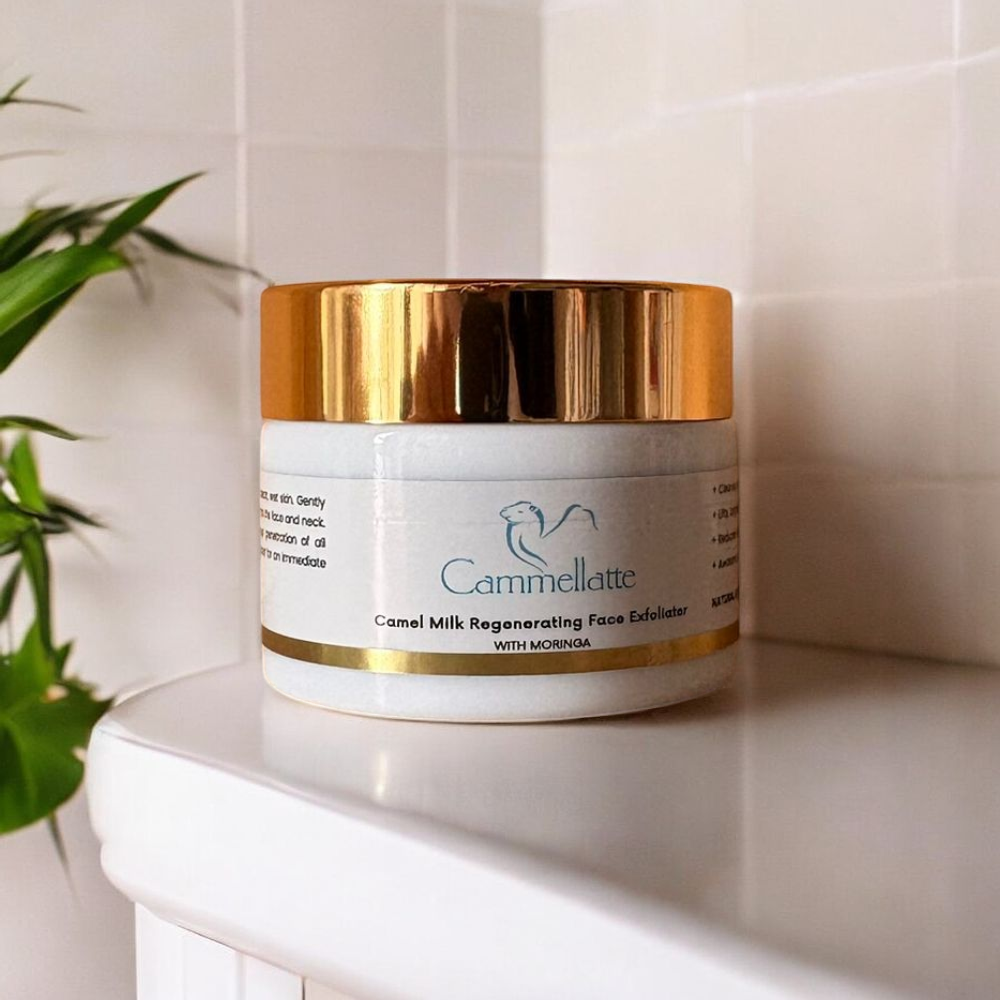 A jar of Cammellattes Camel Milk Regenerating Face Exfoliator with Moringa, featuring a white body and gold lid, rests on a white surface. Light-colored tiles and a small green plant elegantly enhance the luminous skin promise this product delivers.