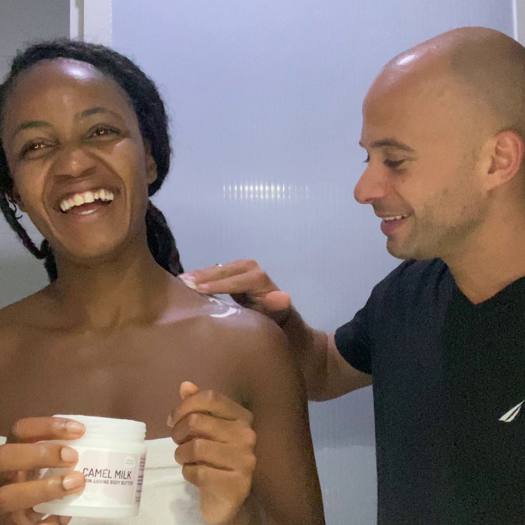 A man in a black shirt applies Cammellattes Delicious Body Butter to a womans shoulder, making her smile at her smooth skin. She is wrapped in a towel in a light room, with the jar nearby.