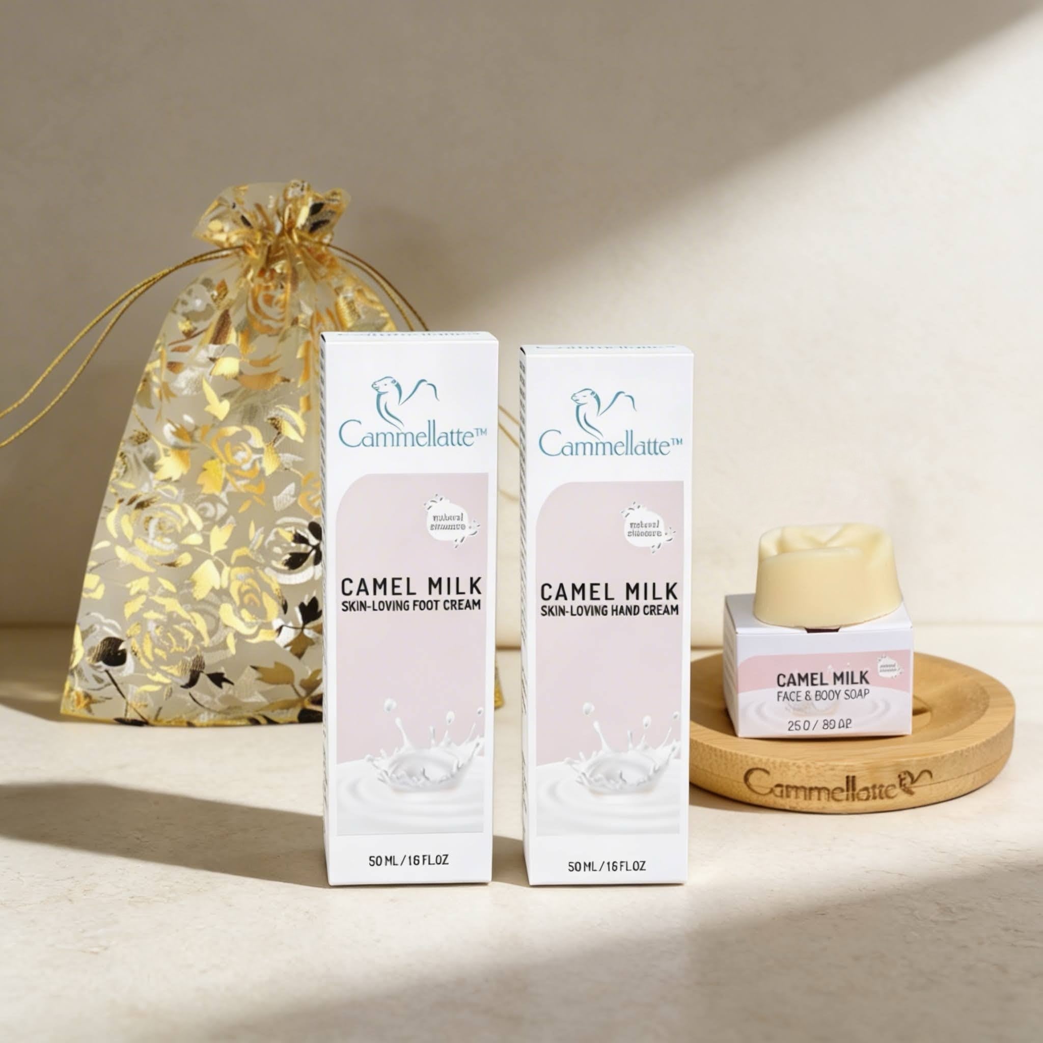 The Cammellatte Hand & Foot Rescue Set, displayed with a gold floral gift bag on a beige surface, includes camel milk skin cream and a small jar of face and body soap arranged on a wooden dish.