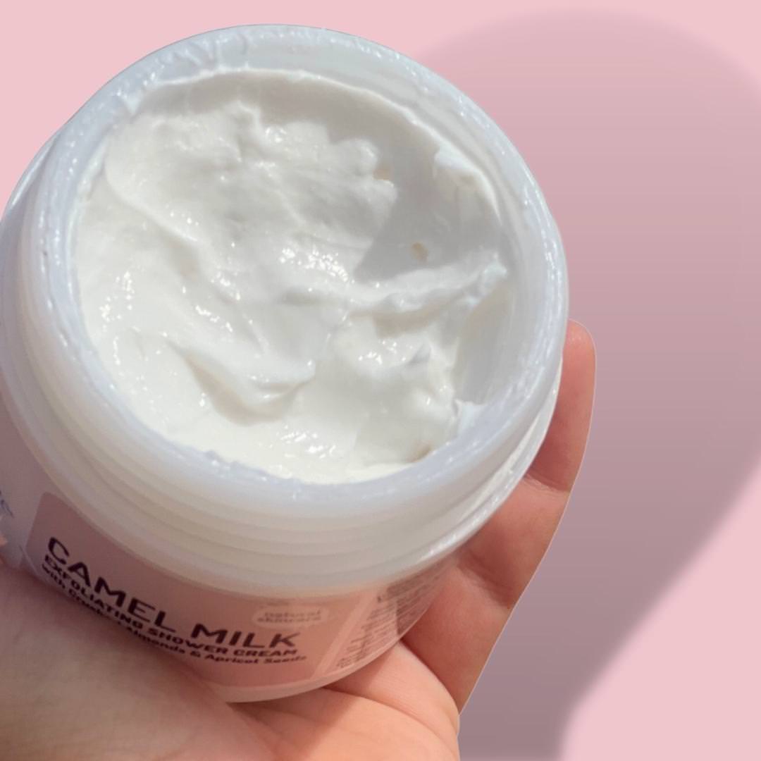 A hand holds an open jar of Cammellatte's luxurious Camel Milk Exfoliating Shower Cream against a pink backdrop, showcasing the smooth, white texture that beautifully complements the label.