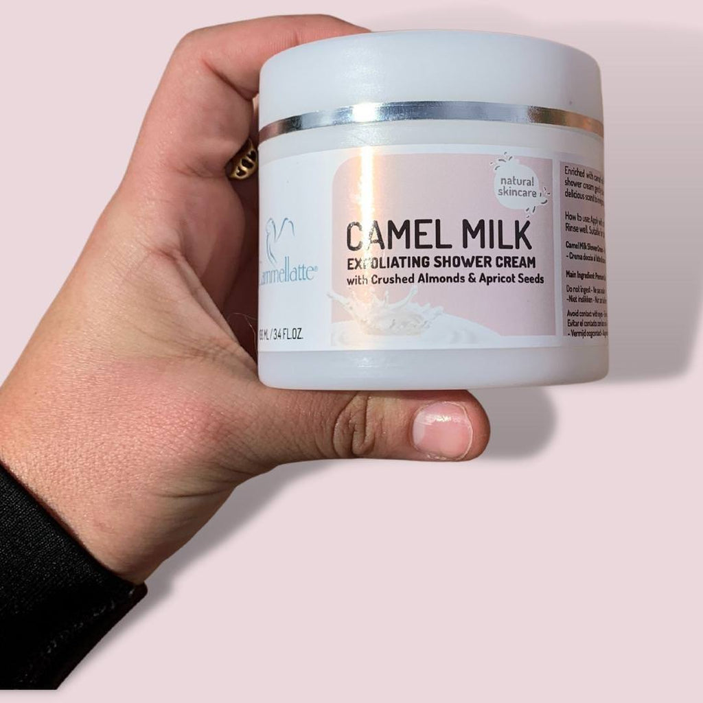 A hand holds a jar of Cammellatte's Camel Milk Exfoliating Shower Cream, featuring ingredients such as crushed almonds and apricot seeds. The light pink background adds a gentle touch to the scene.