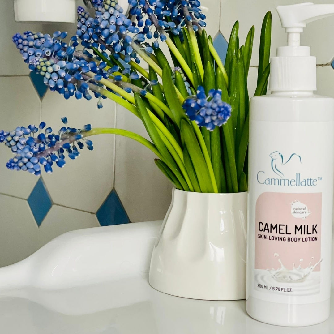 A bottle of Cammellatte Camel Milk Body Lotion, in a pristine white design, sits elegantly on the white sink. Beside it, a vase displays vibrant green stems and clusters of small purple flowers. The backdrop features a striking pattern of white and blue hexagonal tiles.