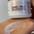 A close-up of a hand highlights a smear of camel milk-infused cream while holding a container labeled 