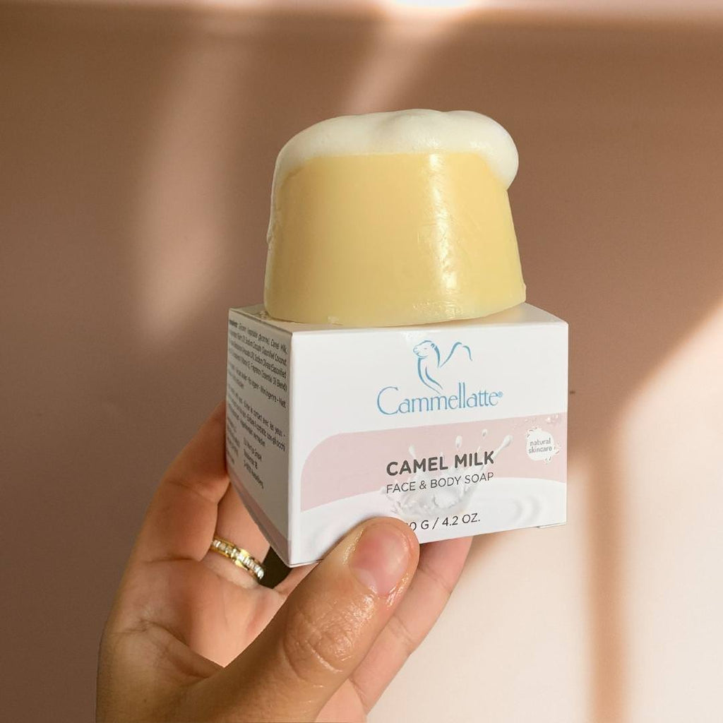 A hand holds a Cammellatte Camel Milk Face & Body Soap, partially unwrapped from its elegant box. The creamy soap is adorned with a rich, foamy lather. Set against a soft, neutral background, the scene exudes tranquility and luxury.