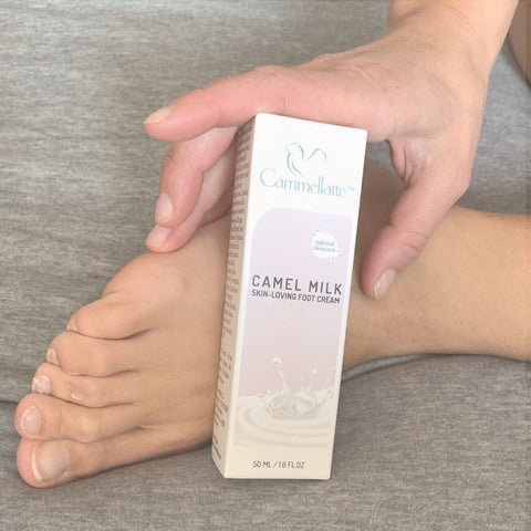 A hand holds a box of Cammellatte Foot Care Bundle beside bare feet on a gray surface, offering a deeply hydrating boost to soothe dry, cracked skin.