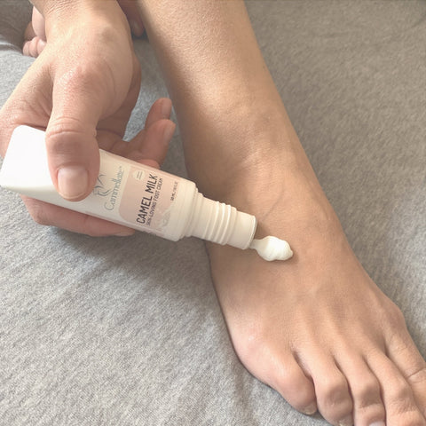 A person applies Cammellattes deeply hydrating Foot Care Bundle onto the top of their bare foot, resting on a gray fabric surface—ideal for soothing dry, cracked feet.